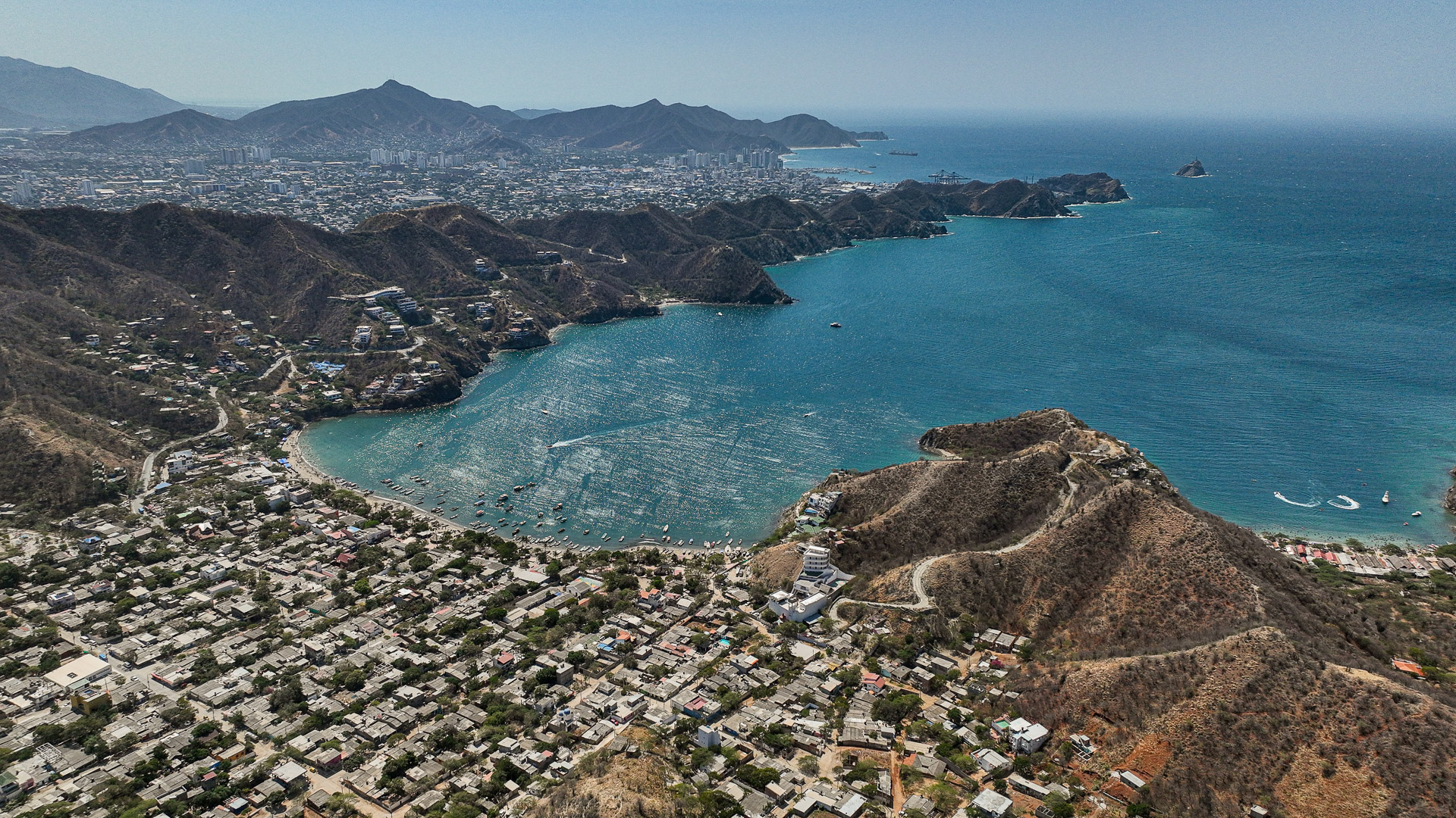 Panoramski pogled z dronom na zaliv Taganga in mesto Santa Marta ob karibski obali Kolumbije, z griči in modrim morjem.